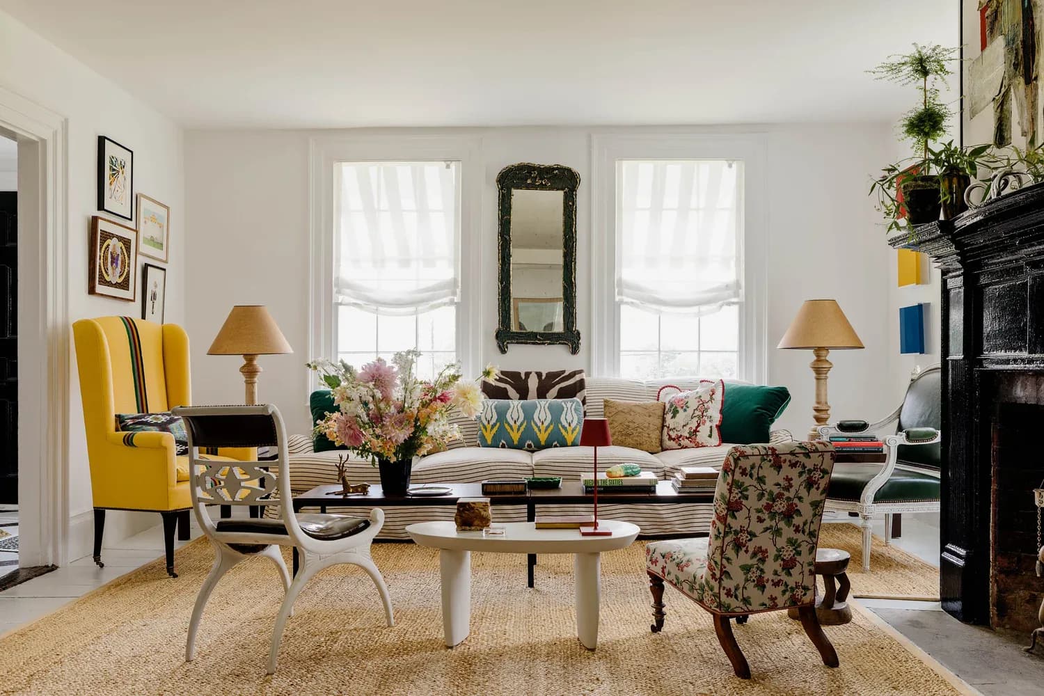 A bright living room features a cream sofa with patterned pillows, yellow wingback chair, floral accent chairs, two windows with roman shades, fireplace, and warm lighting, designed by Nick Olsen.