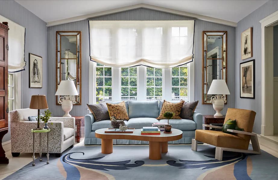 Light-filled living room with blue-gray walls, blue sofa, patterned area rug, wood cocktail table, and brass-framed mirrors flanking tall windows by Nick Olsen.