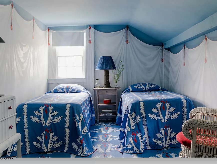 A twin bedroom with blue ceilings and draped white fabric canopies, two navy patterned beds, and traditional furnishings designed by Nick Olsen.