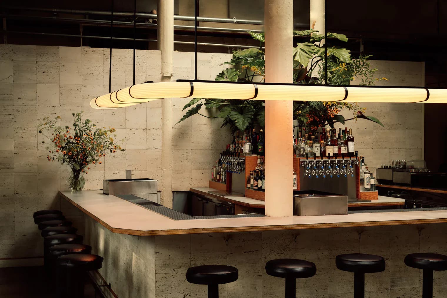 Modern bar interior with curved linear pendant lighting, white brick walls, potted plants, and black bar stools by Nick Ozemba.
