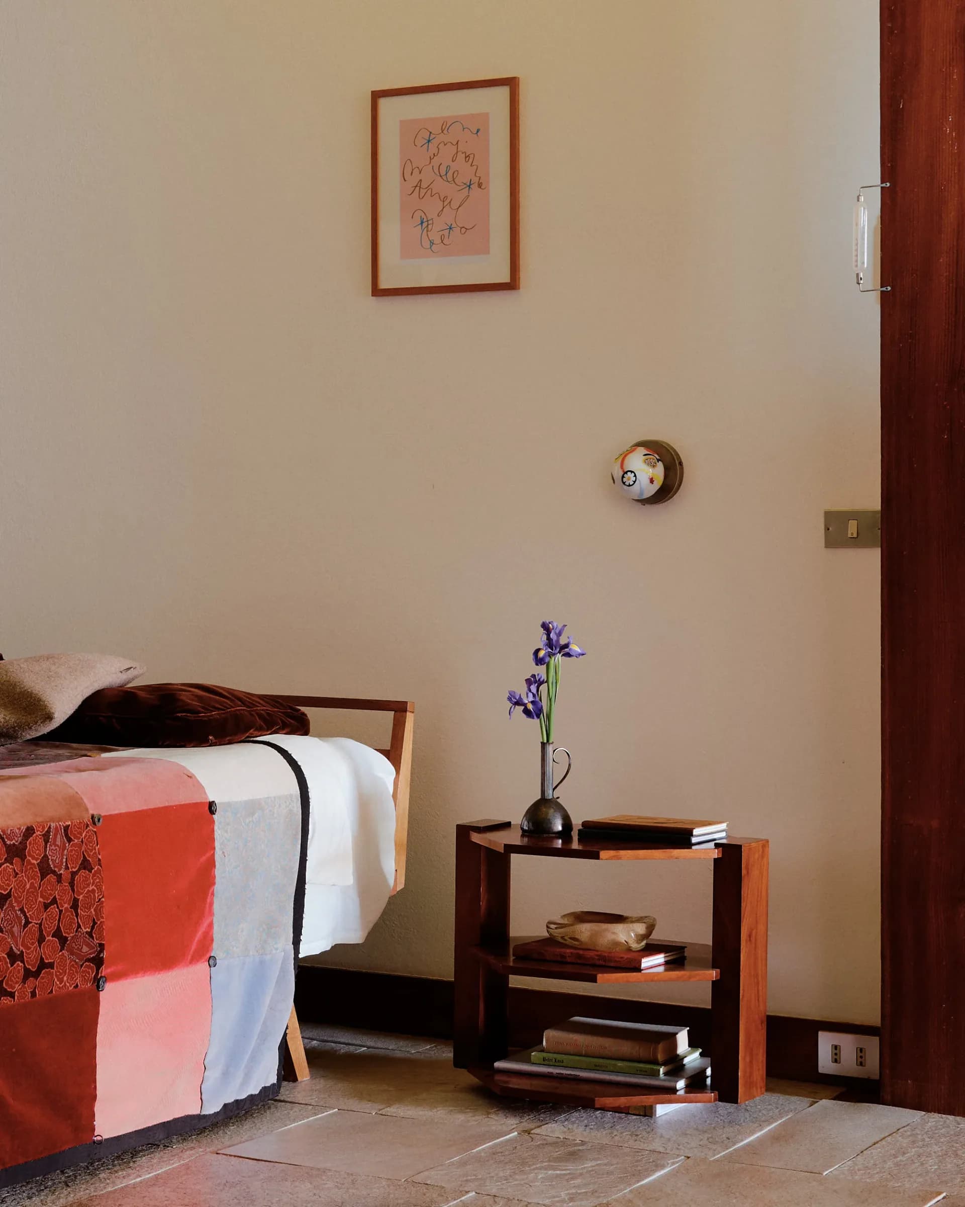 Minimalist bedroom with warm beige walls, wooden bed frame, dark wood nightstand with purple flowers, framed sketch above, and round wall clock by Nick Ozemba.