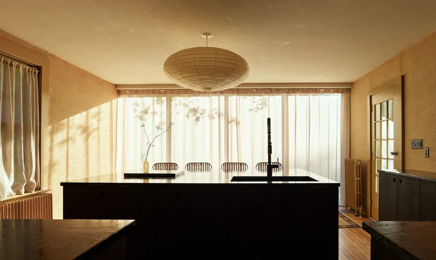 Minimalist kitchen with black island, warm wood tones, and large windows overlooking trees, designed by Nick Spain.