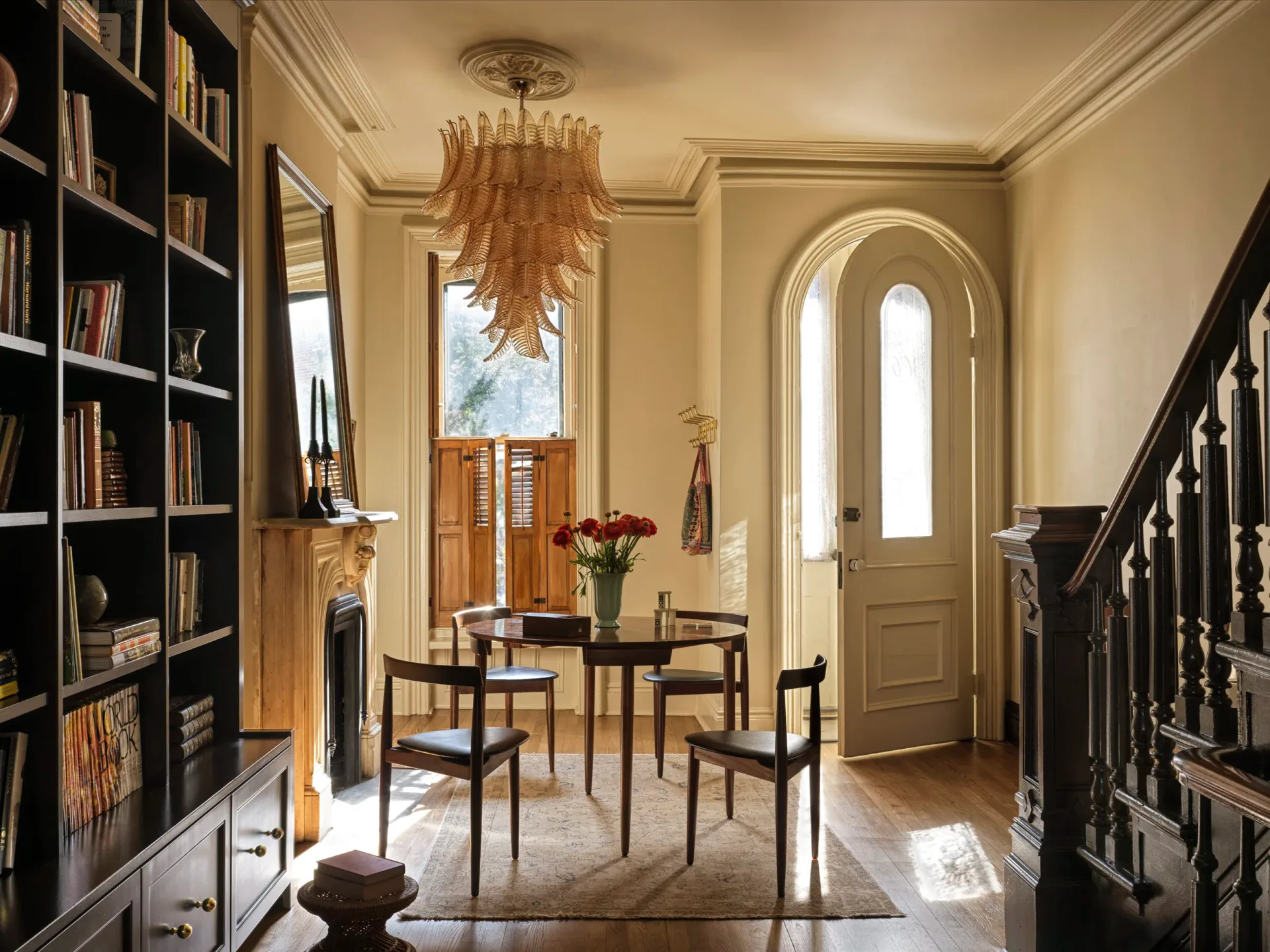 Elegant foyer with cream walls, dark built-in shelving, wooden dining chairs, arched doorway, and brass chandelier by Nick Spain.