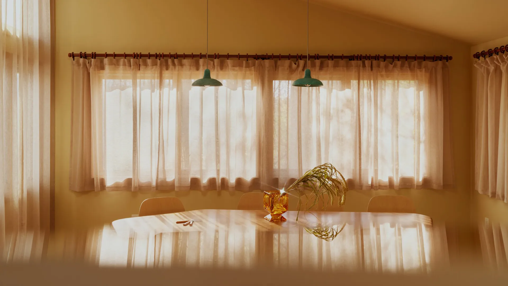 Warm-toned dining room with sheer curtains filtering golden light, pendant lamps, and minimalist furnishings by Nick Spain.