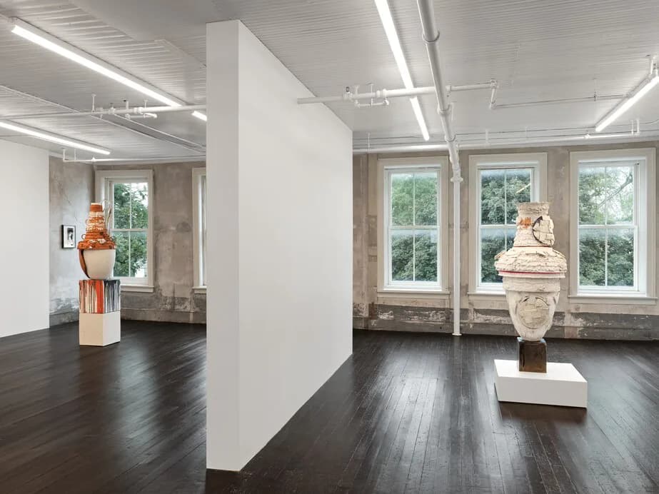 A minimalist gallery interior with dark wood flooring, white partition walls, large windows, and sculptural busts on pedestals, rendered in crisp synthetic lighting by Nicole Cherubini.