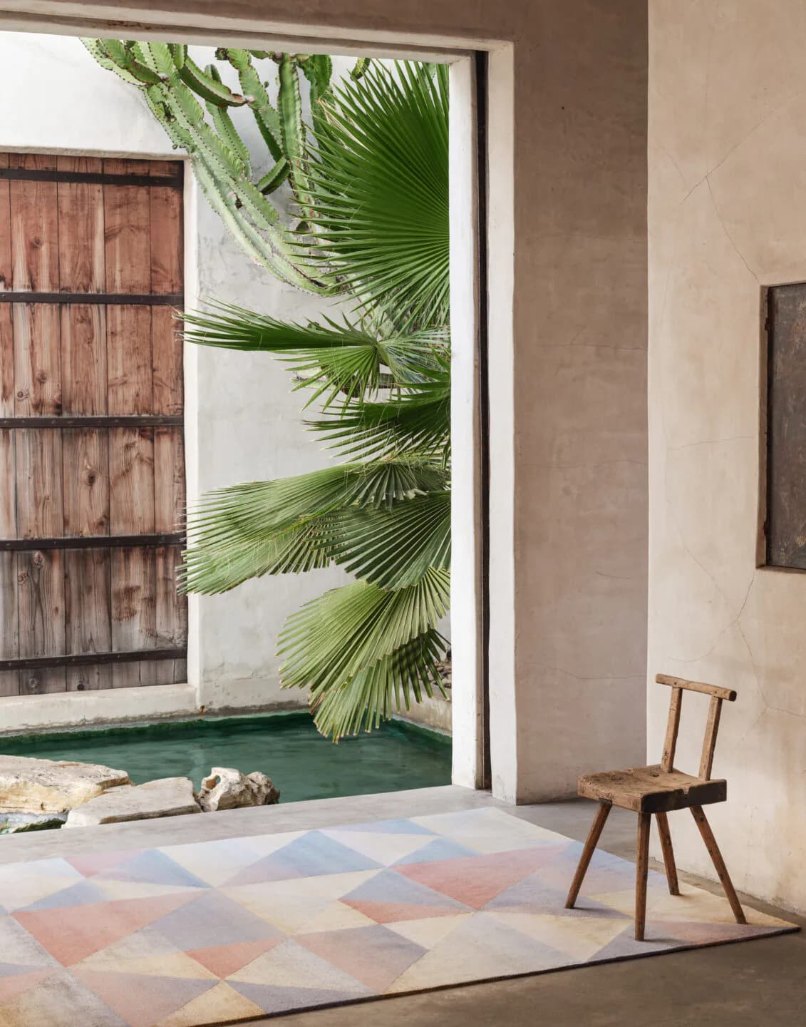 Minimalist interior with geometric pastel rug, wooden chair, and palm-framed courtyard view by Nicole Fuller.