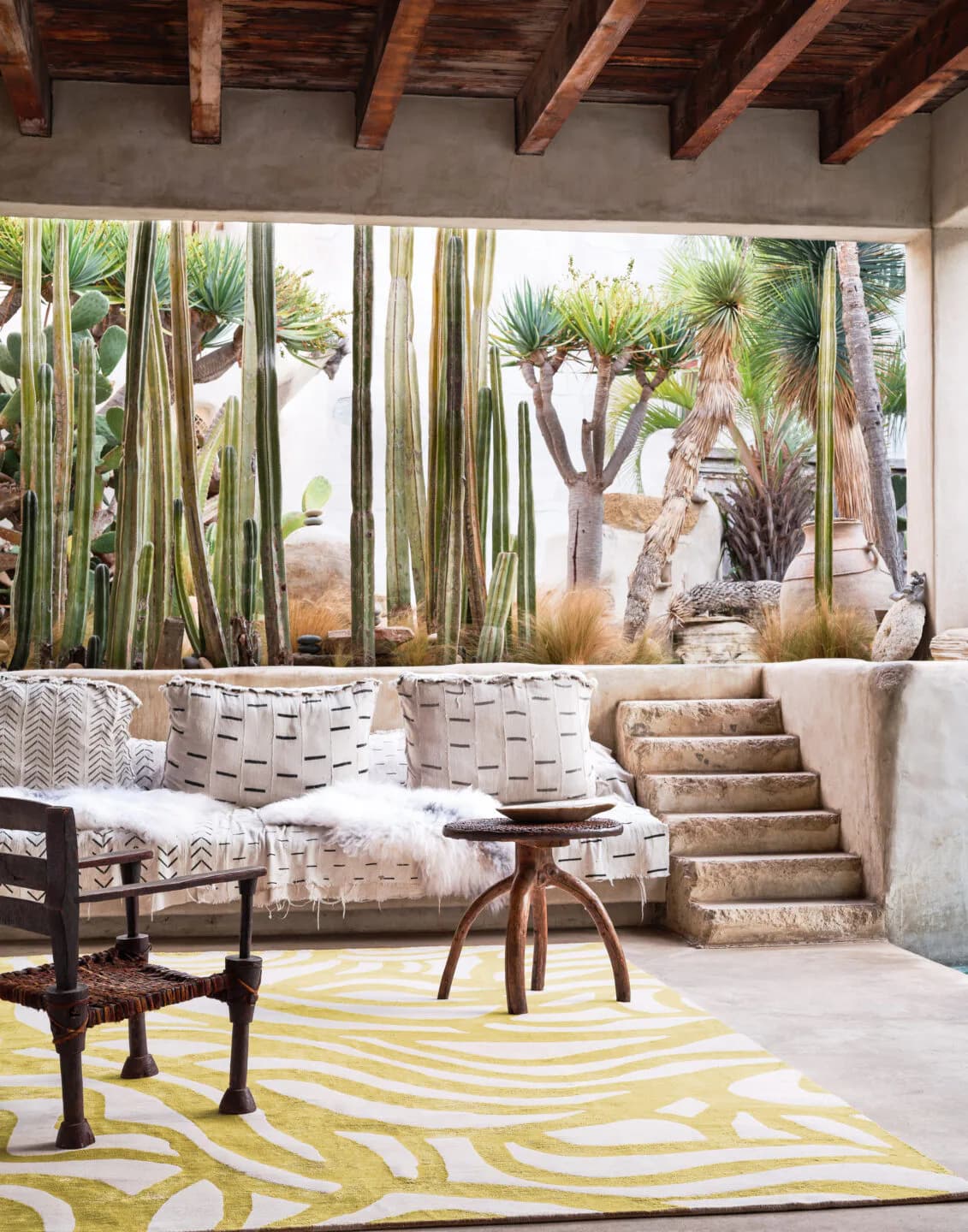 Covered patio with exposed wooden beams, built-in seating with patterned cushions, yellow striped rug, and cacti garden views by Nicole Fuller.