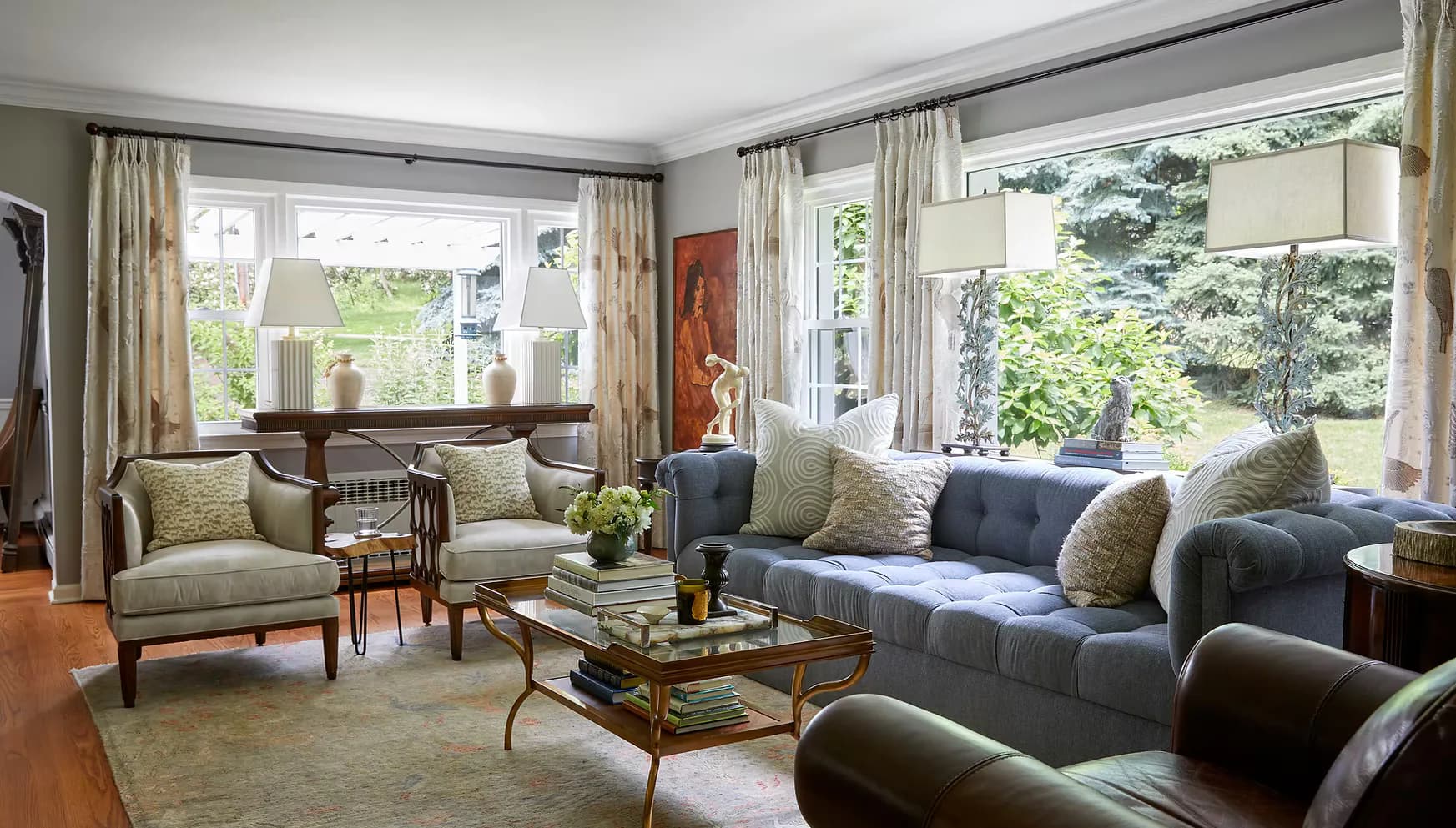 Elegant living room with neutral upholstered seating, mid-century modern furniture, large windows overlooking gardens, soft natural light, and layered textiles in greens and neutrals by Nicole Peter.