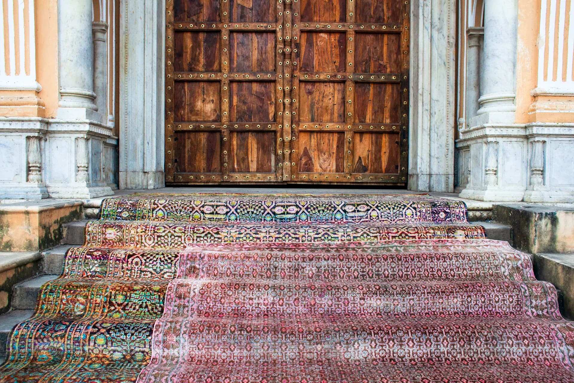 Ornate entry foyer featuring vibrant patterned runner carpets in jewel tones leading to weathered wooden doors flanked by classical columns, designed by Nicole Pierpont.