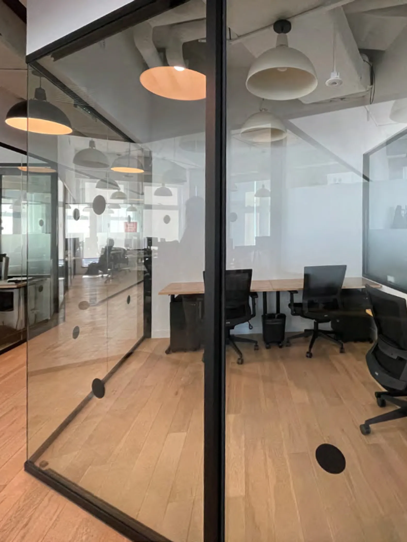 Modern office space with glass-partitioned rooms, wooden flooring, pendant lighting, and black office chairs, designed by Nicole Robertson.