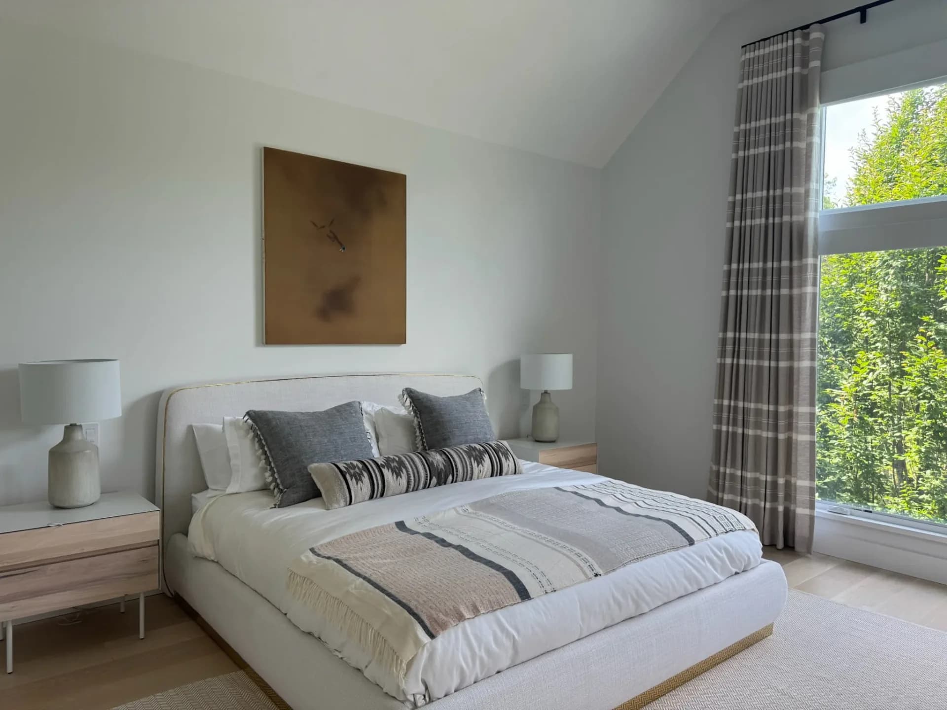 Serene bedroom with cream upholstered bed, warm wood nightstands, layered bedding, abstract bronze artwork, and plaid curtains framing large windows by Nina Fisher.
