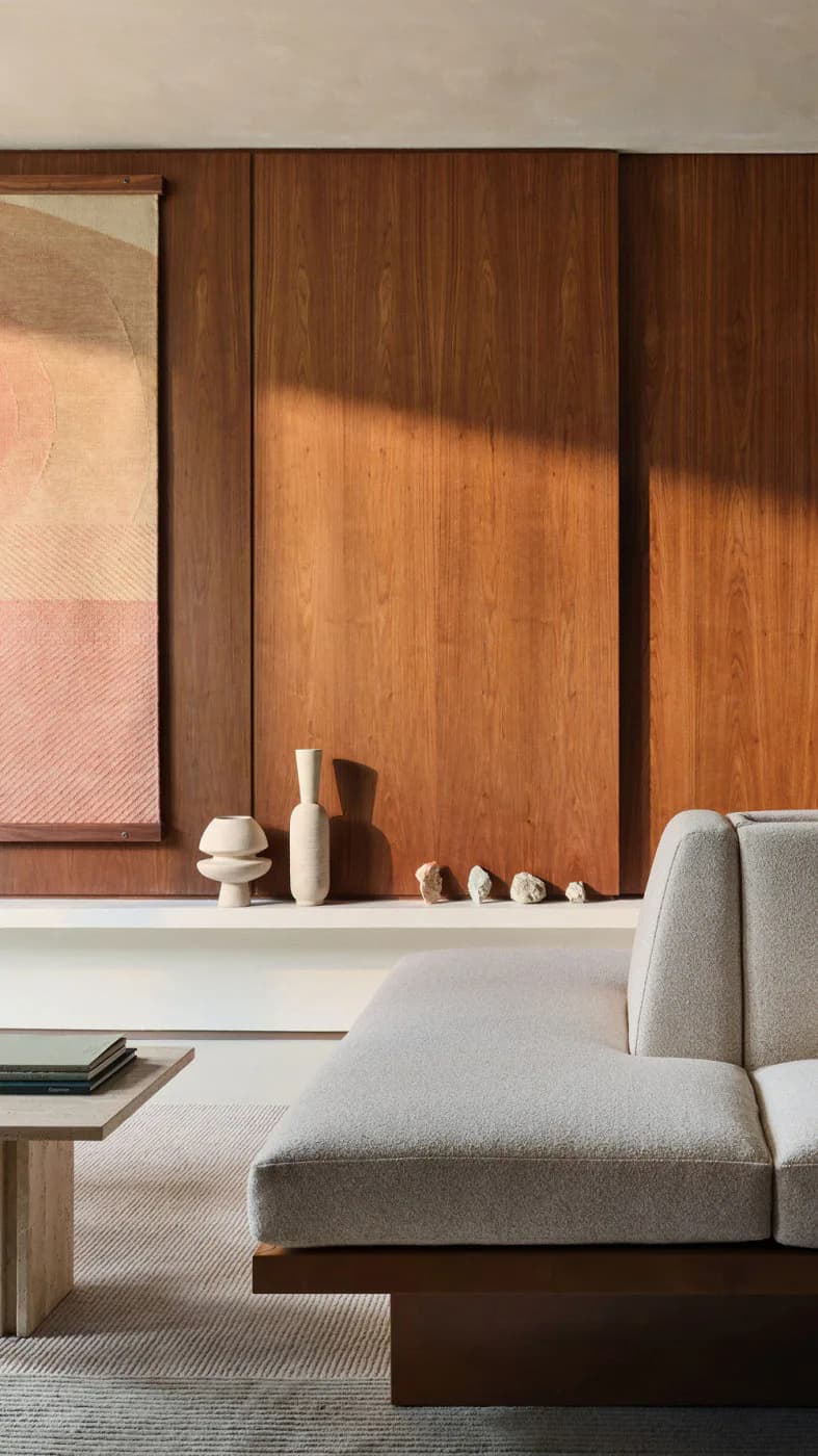 Modern living room featuring warm wood-paneled wall with artwork, light gray upholstered sofa, minimalist shelf with ceramic objects, and natural light creating geometric shadows, by Nina Jakic.