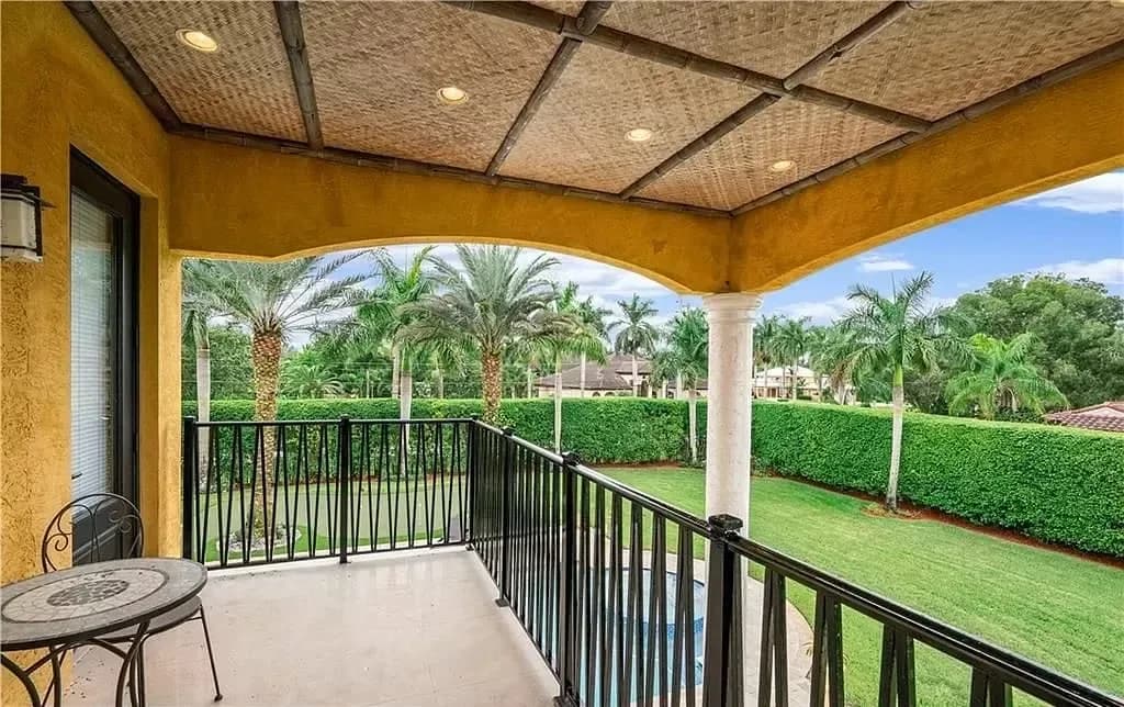 Covered balcony with yellow ochre walls, black metal railings, and woven ceiling detail overlooking manicured lawn and palm trees by Nina Nielsen-Norwood.