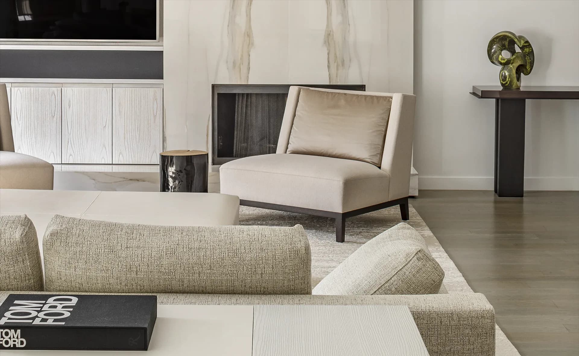 Modern living room with beige sectional sofa, cream accent chair, dark wood shelving, and polished concrete flooring in a minimalist palette by Nitzan Tagansky.