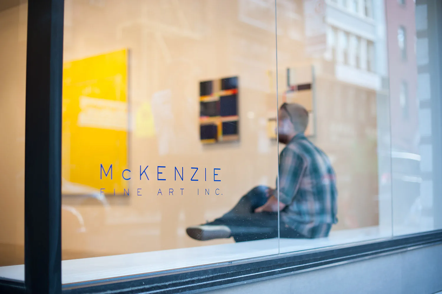 McKenzie Fine Art gallery storefront with yellow signage, abstract artwork on cream walls, and figure visible through glass by Niyati Singh.