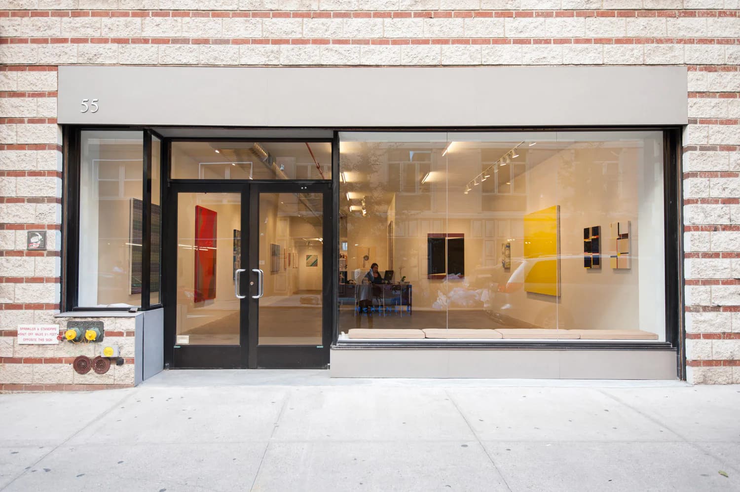 Modern gallery storefront with brick facade, floor-to-ceiling glass windows, black frame entry door, and minimalist interior displaying colorful artwork installations by Niyati Singh.