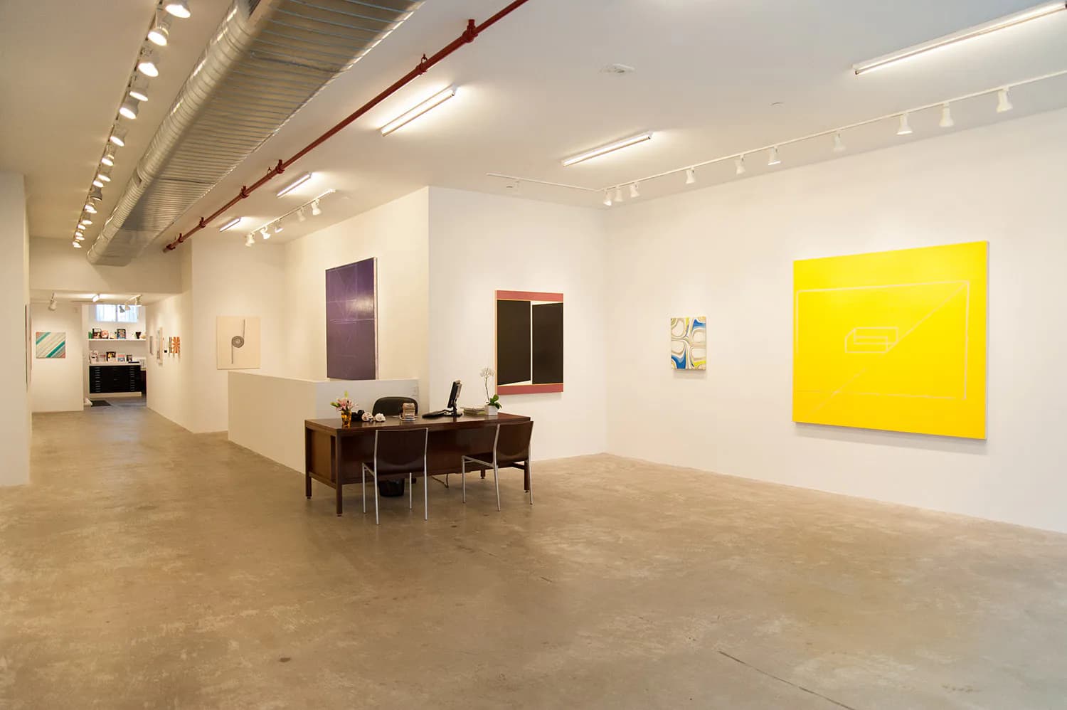 Modern gallery space with polished concrete floors, white walls, and bright yellow abstract artwork, featuring mid-century furniture by Niyati Singh.