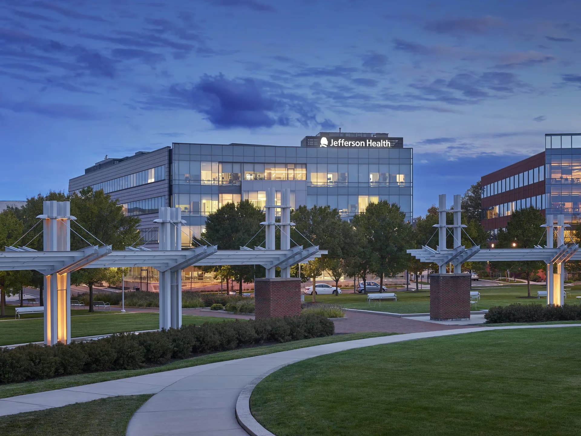Twilight exterior of Jefferson Health medical campus with illuminated modern building, landscaped plaza, curved walkways, and decorative columns by Noah Tolson.