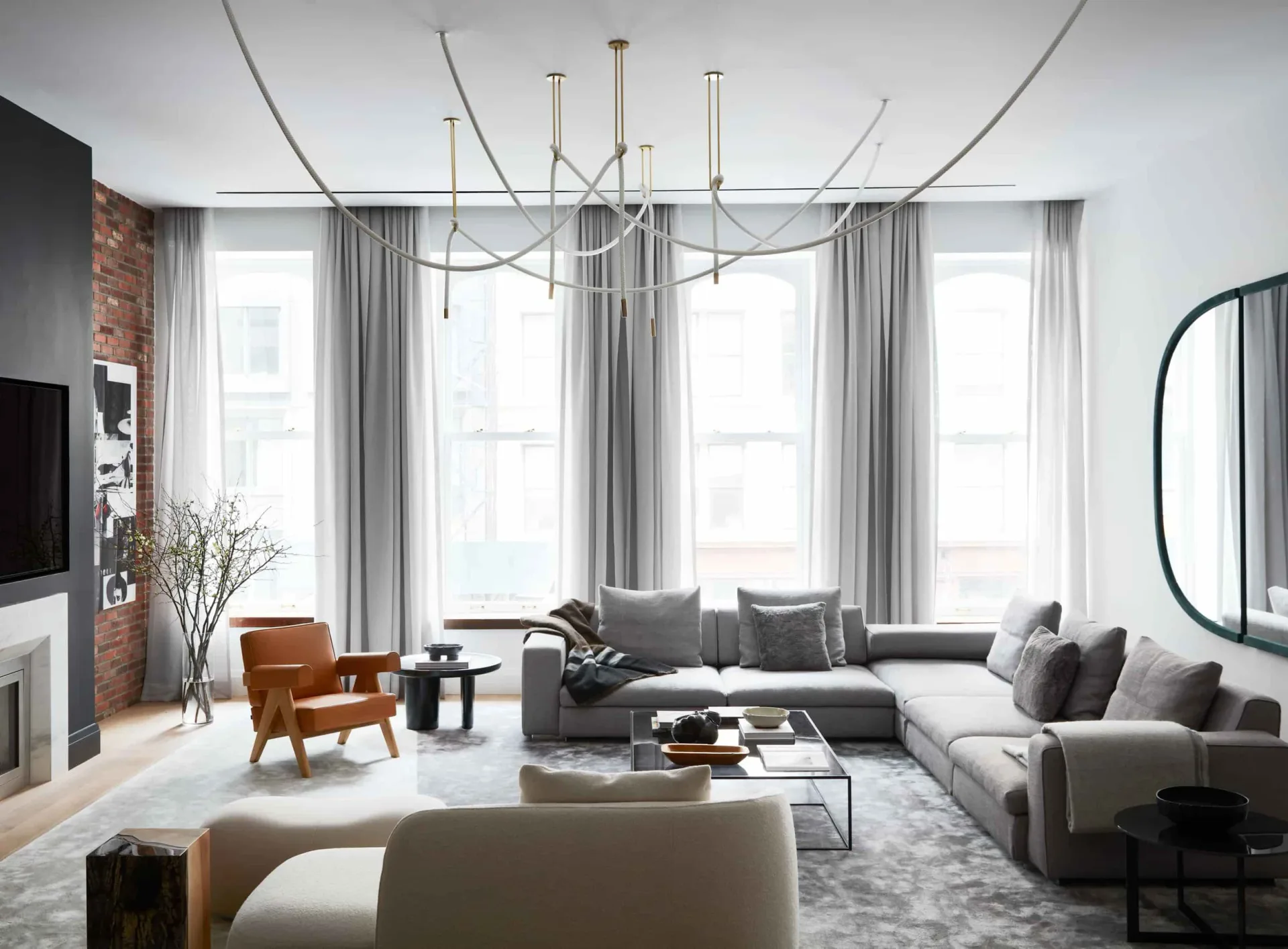 Modern living room with gray sectional sofa, sculptural pendant lighting, floor-to-ceiling windows with gray curtains, and mixed materials including brick and brass accents by Noha Hassan.