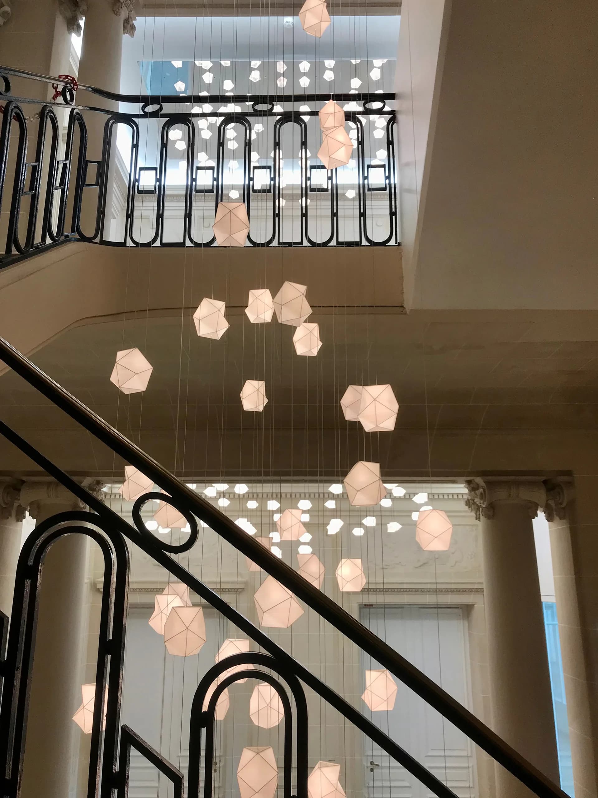 Multi-story atrium with black metal railings and suspended geometric paper light fixtures in soft pink tones, designed by Octavio Amado.