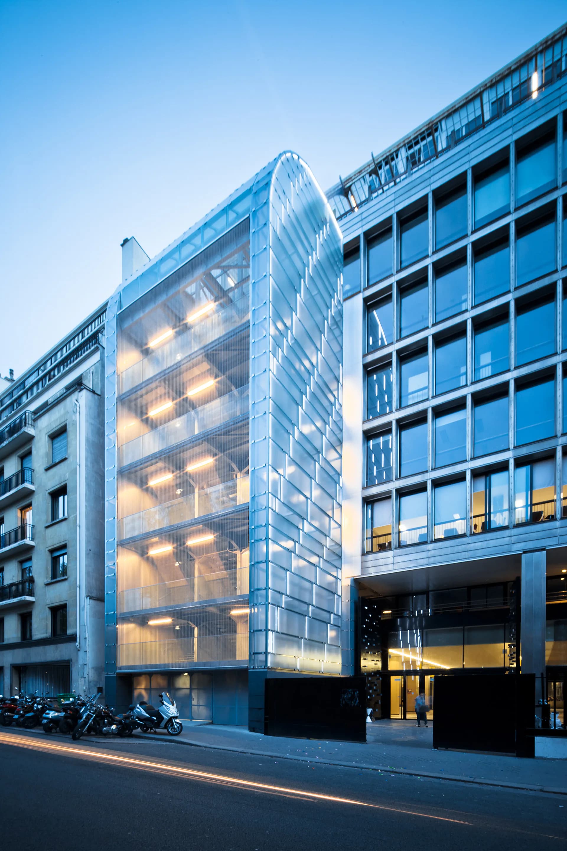 Modern glass-fronted building with illuminated interior at dusk, featuring translucent façade beside traditional architecture by Octavio Amado.