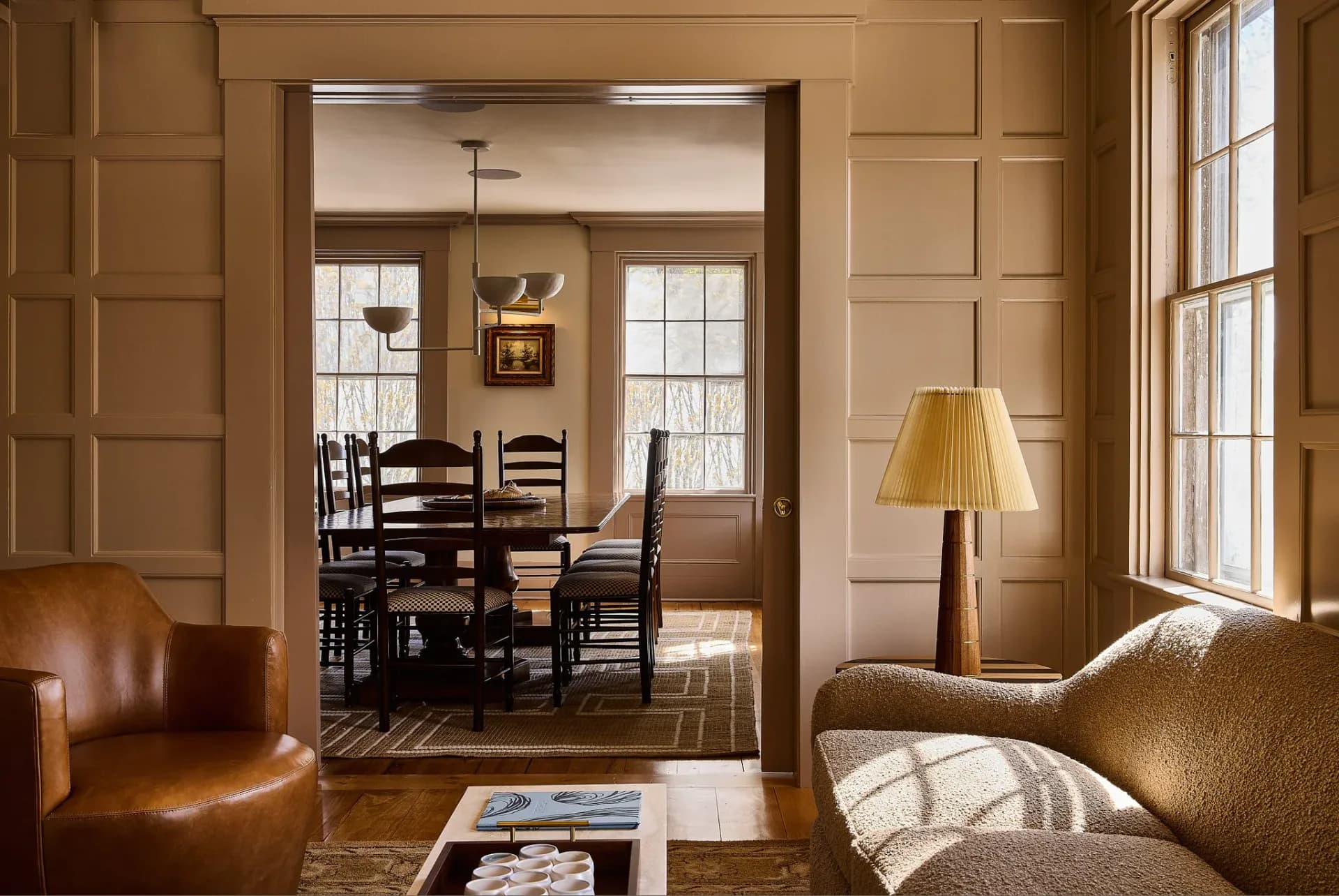 Warm-toned living room with leather seating, natural light from tall windows, and views into a dining area with ladder-back chairs, designed by Oliver Haslegrave.