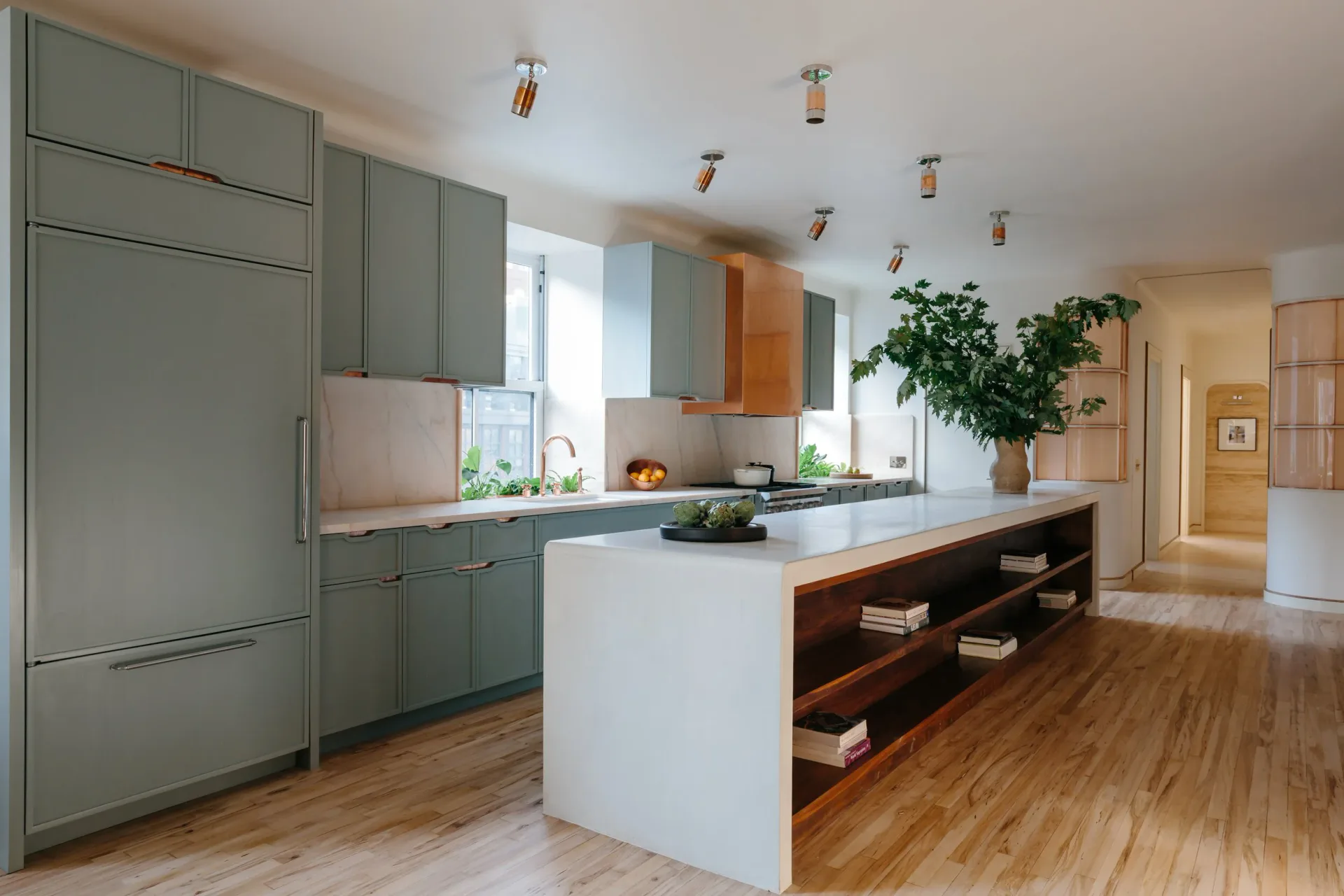 Modern kitchen with sage green cabinetry, white waterfall island with wood shelving, oak flooring, and pendant lighting by Oliver Haslegrave.