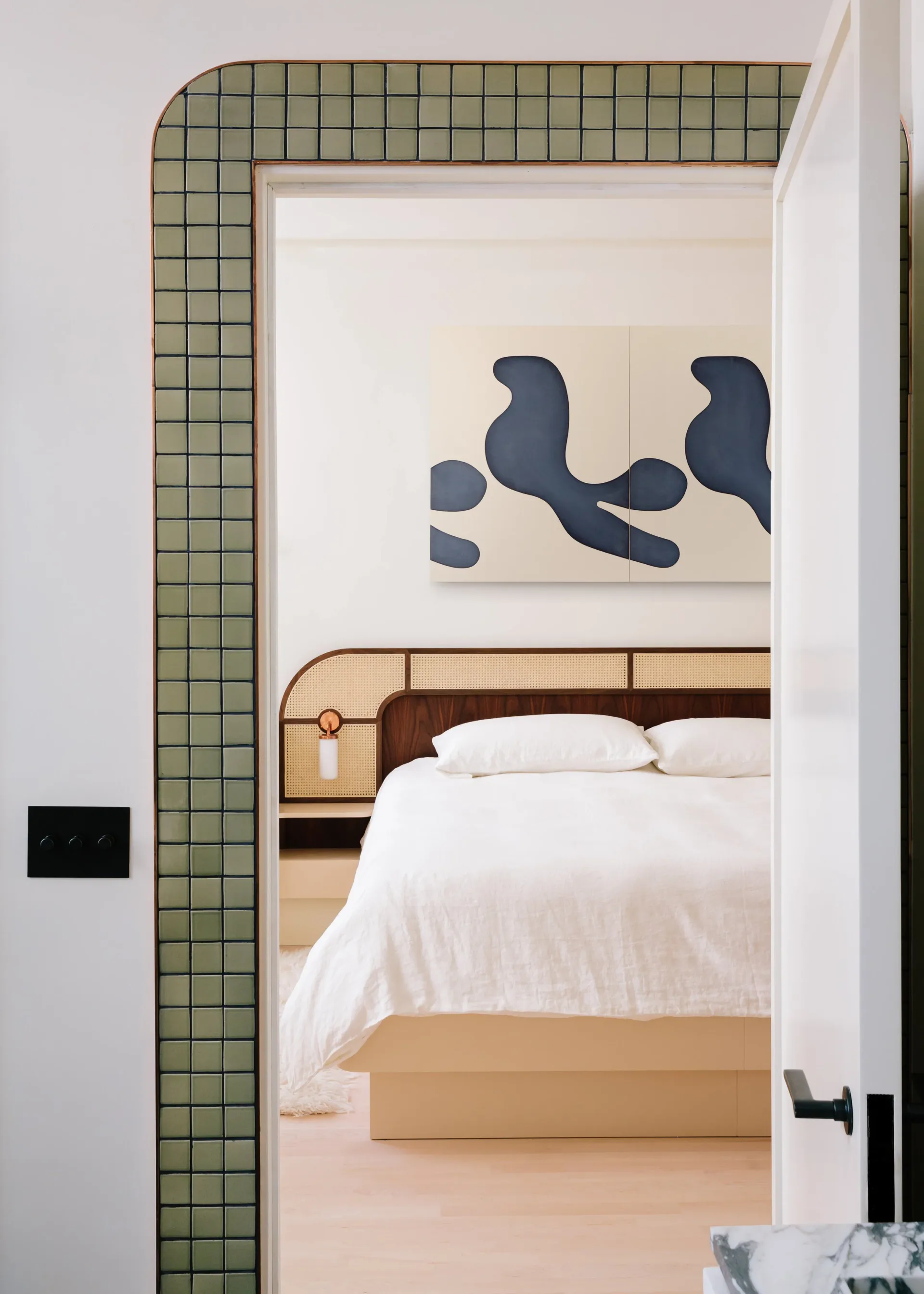 Bedroom with sage green tiled accent wall framing doorway, white bedding, wooden headboard, and abstract dark blue artwork by Oliver Haslegrave.