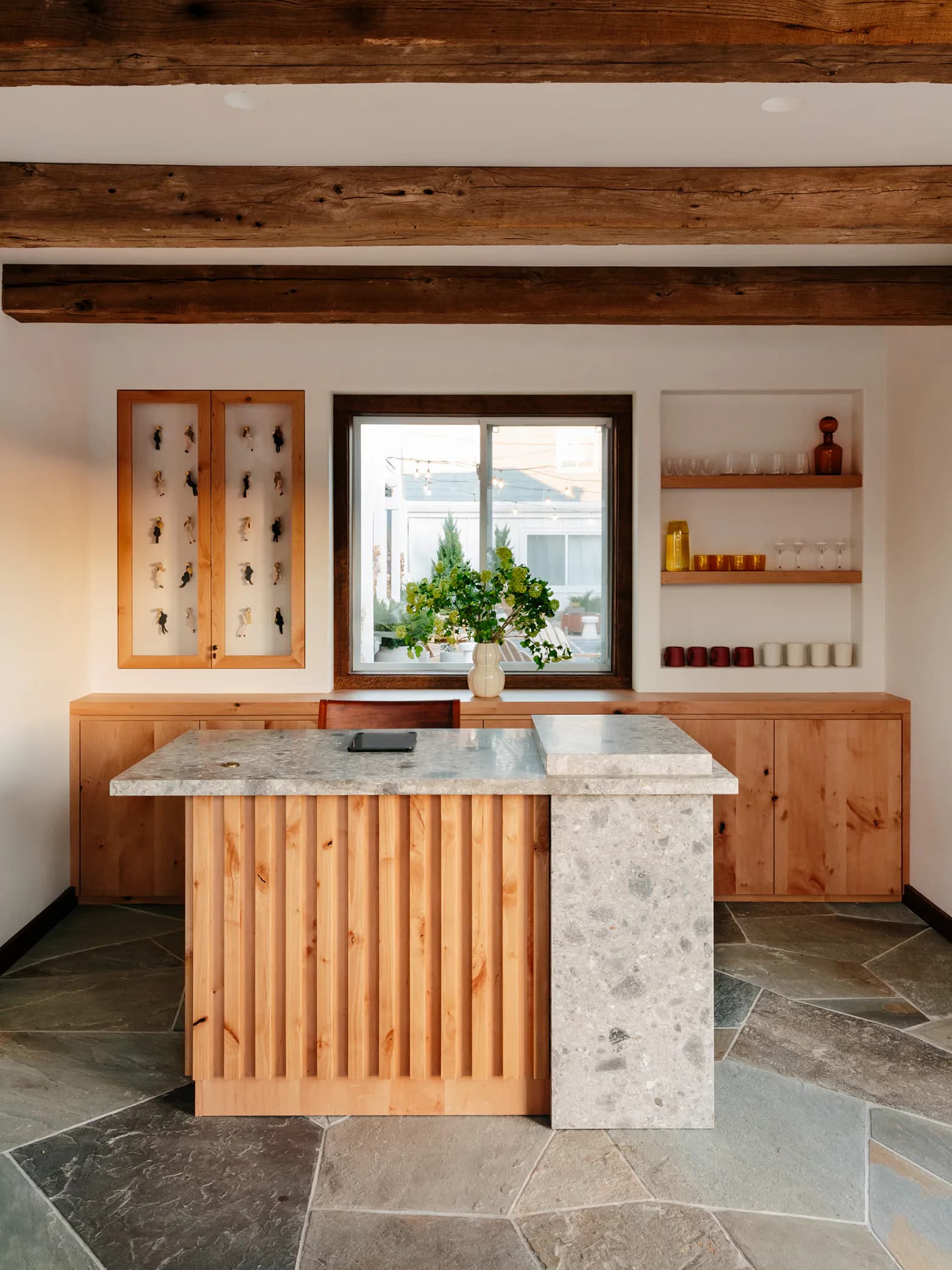 A modern kitchen with warm wood cabinetry, marble waterfall island, exposed timber ceiling beams, and large window overlooking gardens by Oliver Haslegrave.