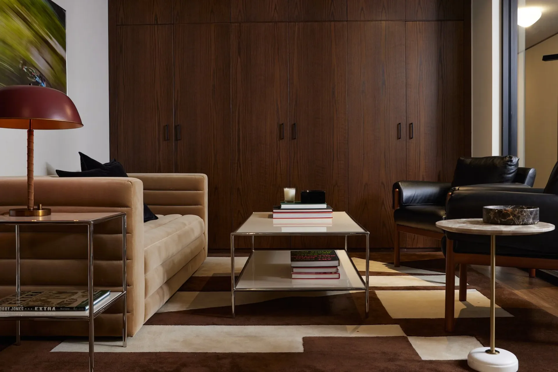 Contemporary living room with dark wood cabinetry, tan upholstered sofa, mixed metal and wood tables, and black accent chairs on geometric area rug by Oliver Rumney.