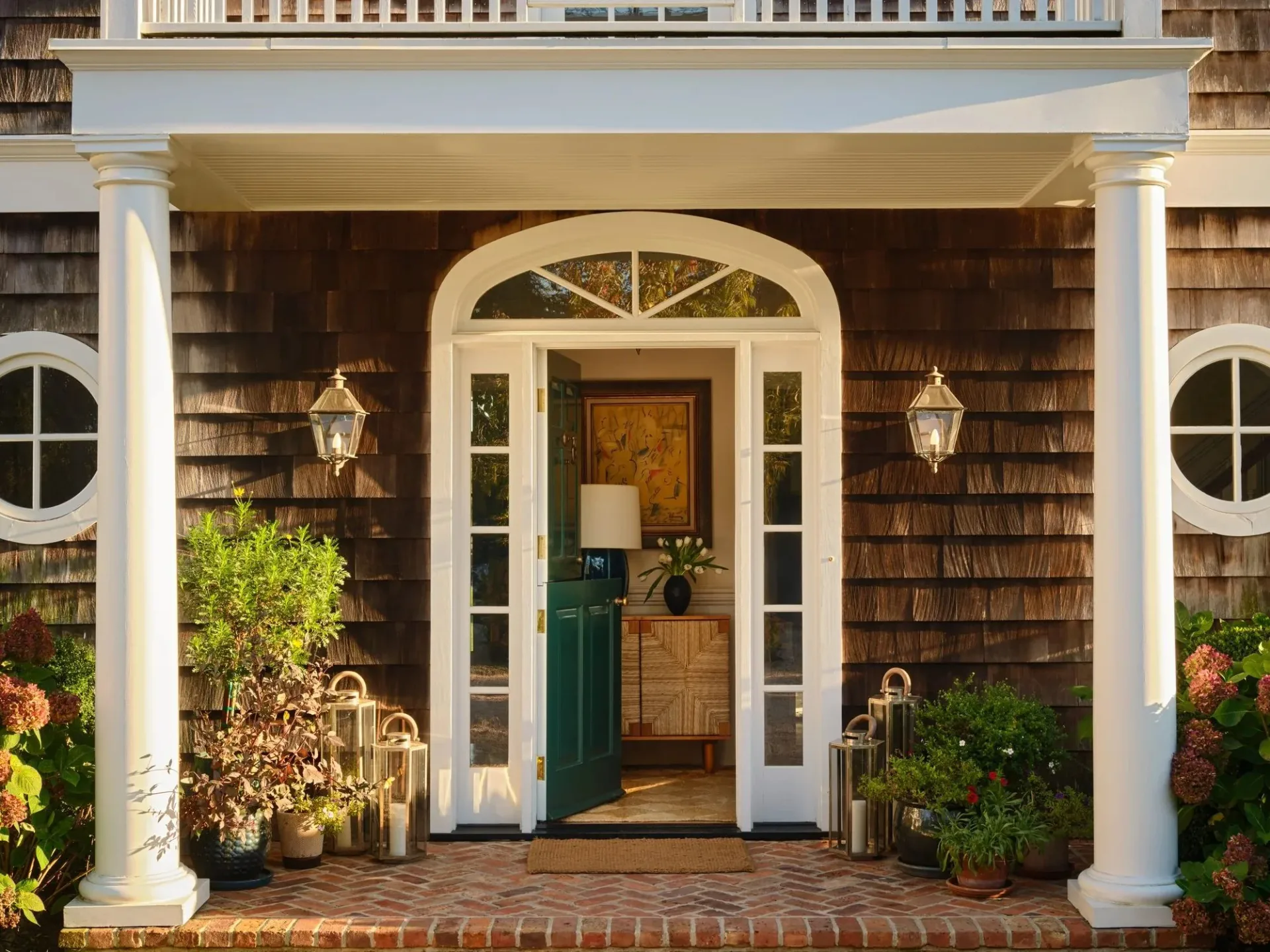 Traditional home entrance with dark wood shingle siding, white columns, arched transom door, symmetrical landscaping, and brass lanterns by Oliver Rumney.