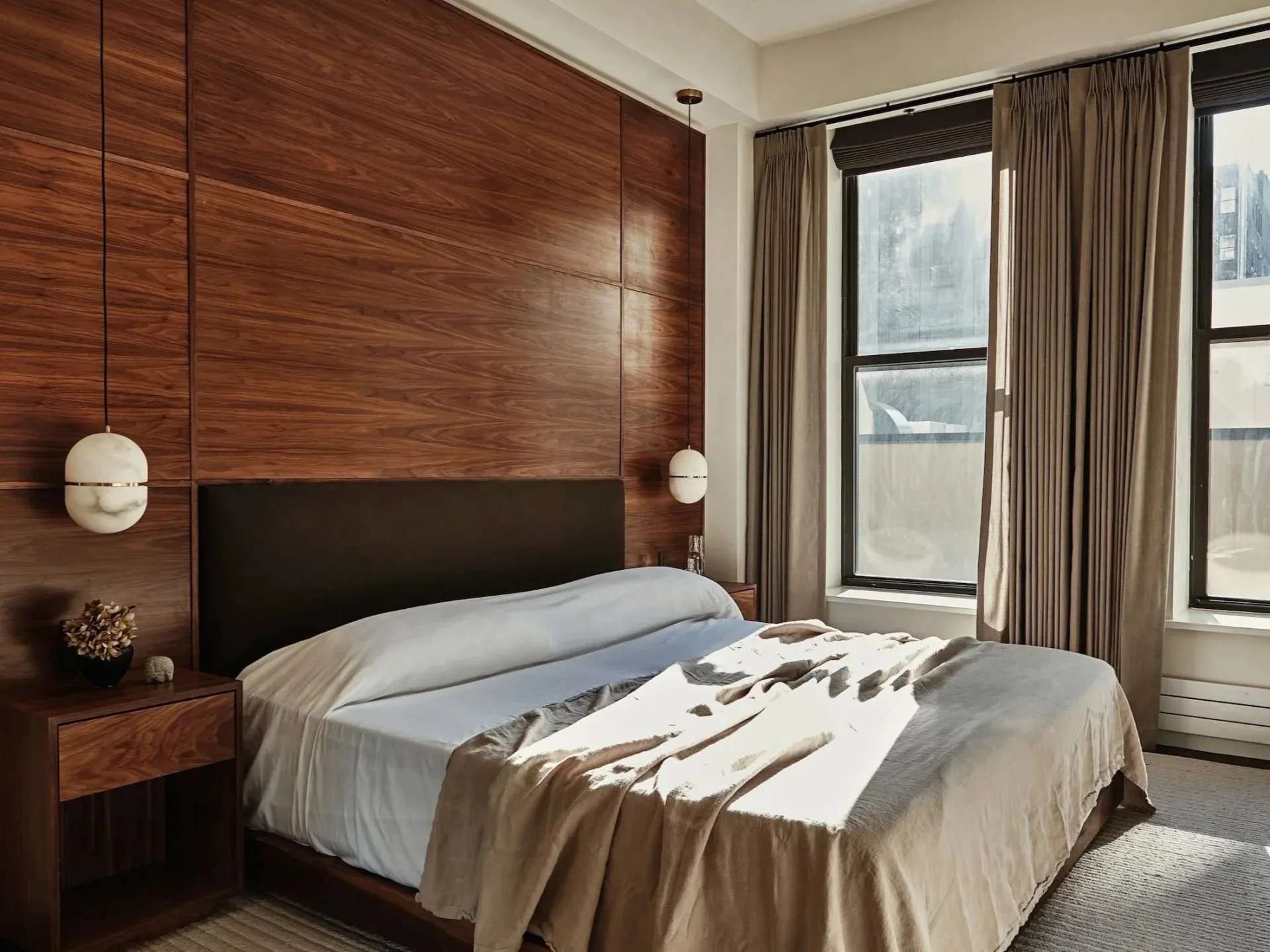 Modern bedroom with rich walnut wood accent wall, black upholstered bed, floating nightstands, and floor-to-ceiling windows with taupe curtains by Oliver Rumney.