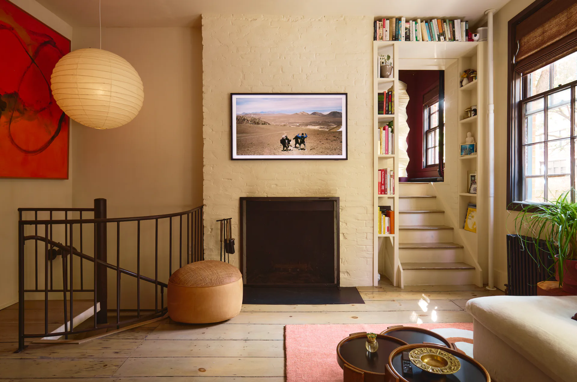 A stylized interior rendering featuring a multi-level loft space with warm wood flooring, cream walls, a red accent wall, floating bookshelves, and carefully composed furnishings in earth tones by Olivia Barry.