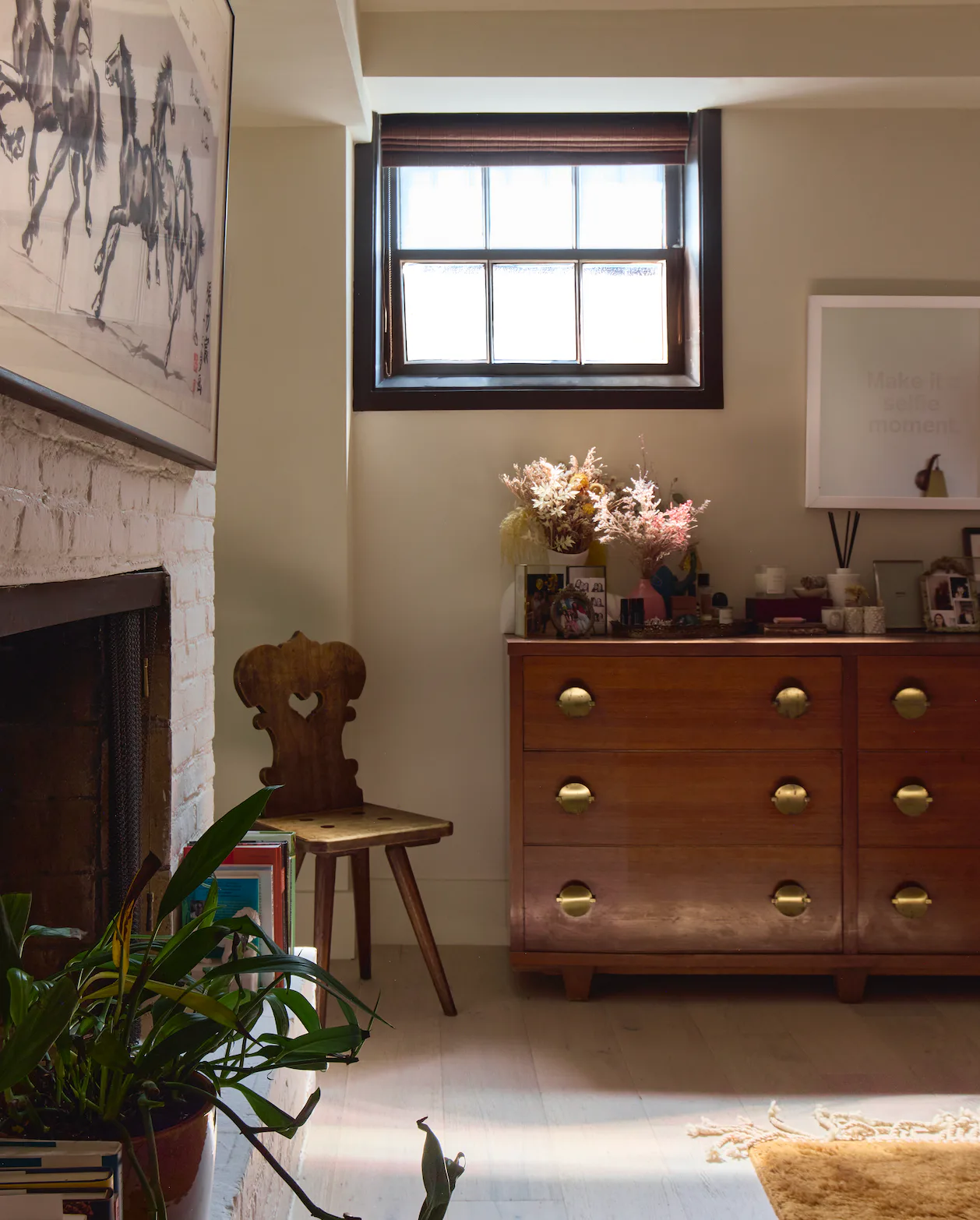 Warm neutral interior with wood dresser, fireplace, potted plant, wooden chair with heart cutout, and window, by Olivia Barry.