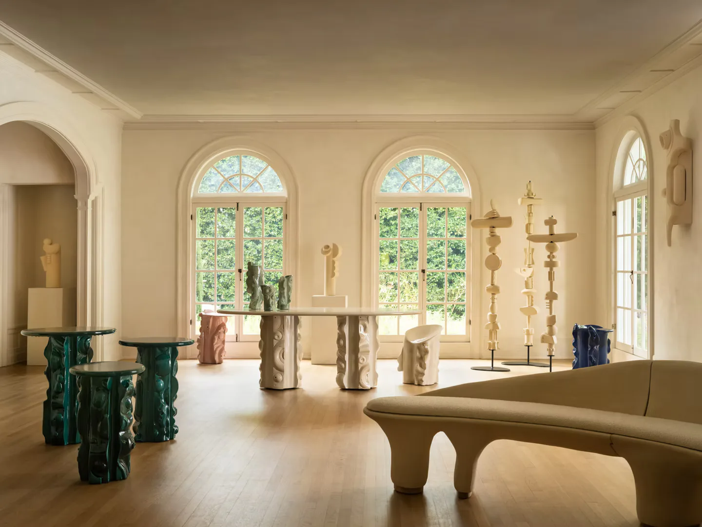 A minimalist chapel interior with cream walls, arched windows, brass candelabras, and sculptural stone pedestals in soft warm lighting by Olivia Cognet.