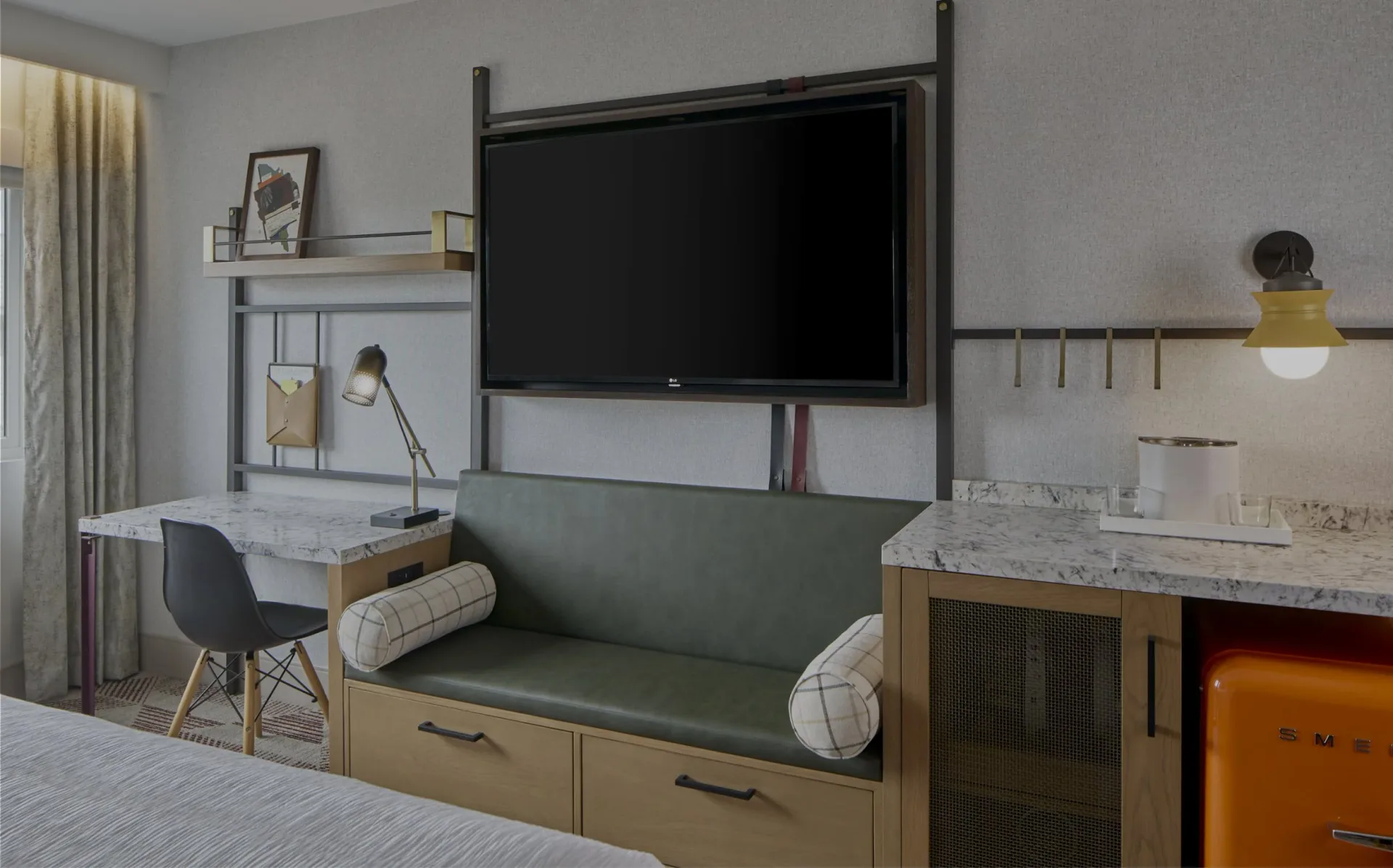 Modern hotel room with integrated TV wall, green banquette seating, wood cabinetry, and marble desk surface designed by Olivia Cooper.