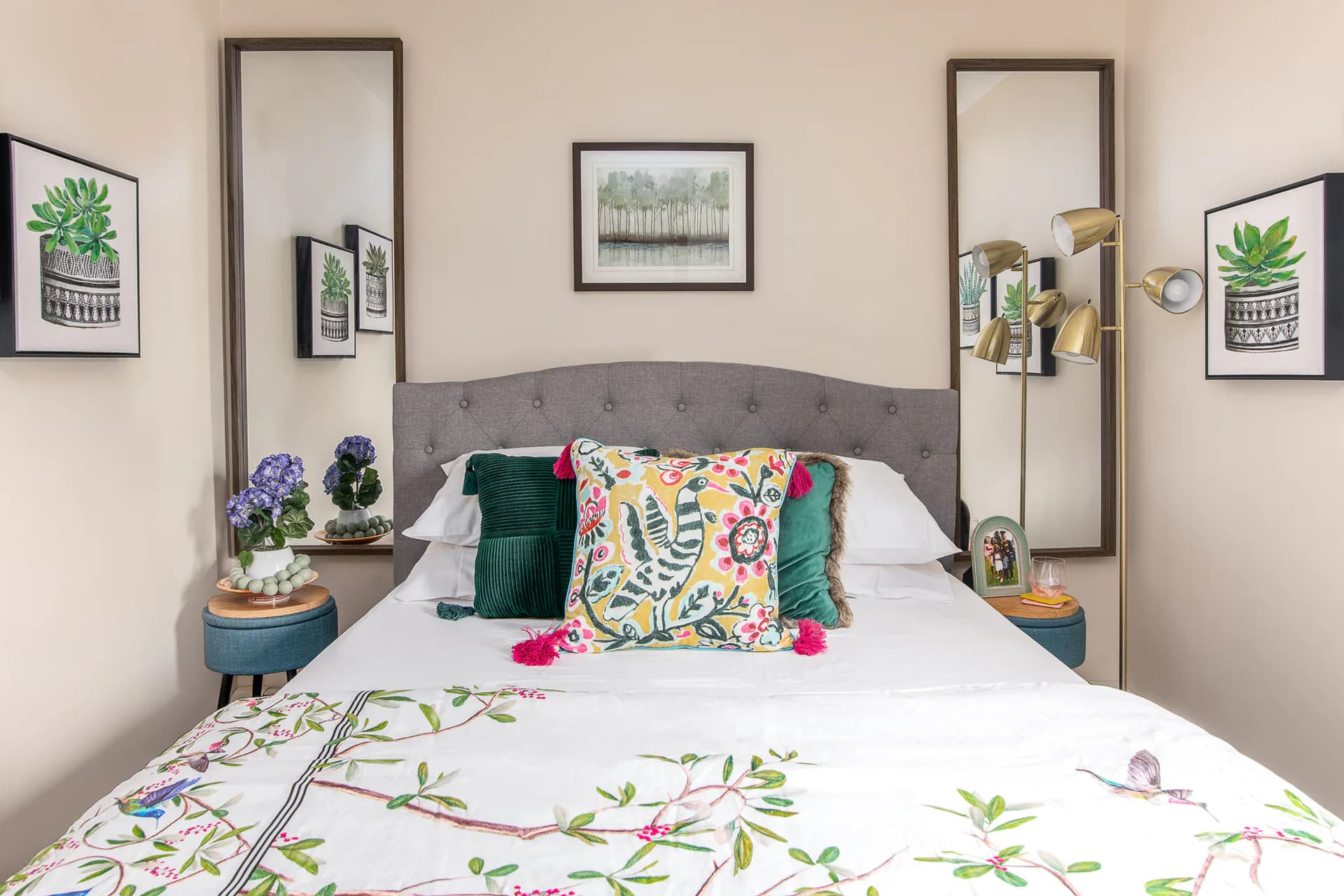 A stylish bedroom features a gray upholstered headboard with built-in shelving, white bedding with botanical prints, colorful accent pillows, and sage-green nightstands, designed by Oreoluwa Oderanti.