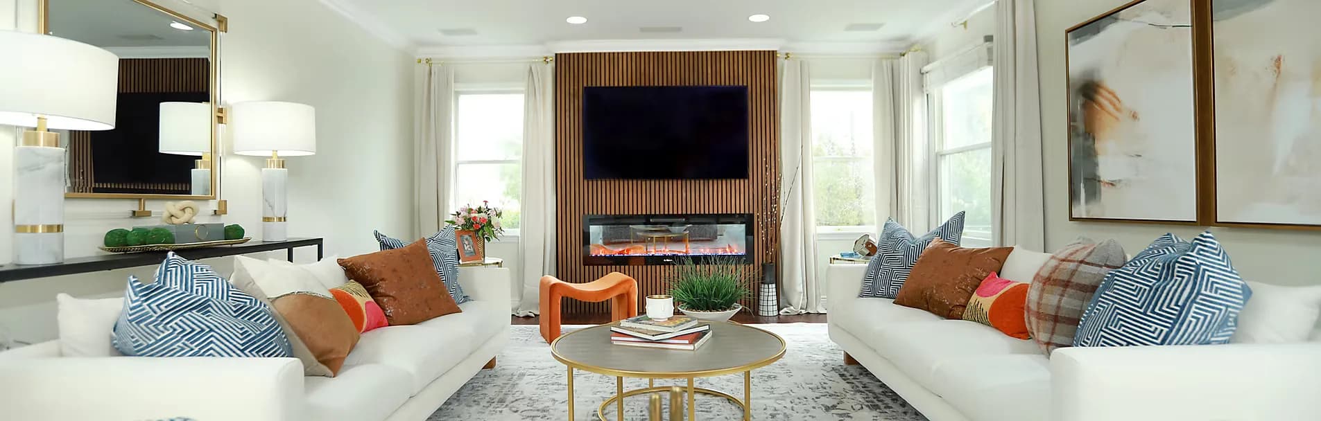 Bright living room with two white sofas, orange and blue accent pillows, fireplace with mounted TV, and neutral palette by Oreoluwa Oderanti.