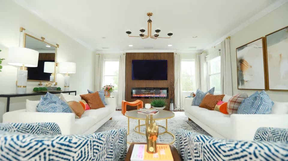 Bright open living room with two white sofas, blue geometric patterned area rugs, orange and blue accent pillows, wooden coffee table, and fireplace with TV, designed by Oreoluwa Oderanti.