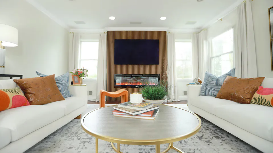 Bright living room with two cream sofas facing a round marble table, wood-paneled TV wall, and warm brown and blue accents throughout by Oreoluwa Oderanti.