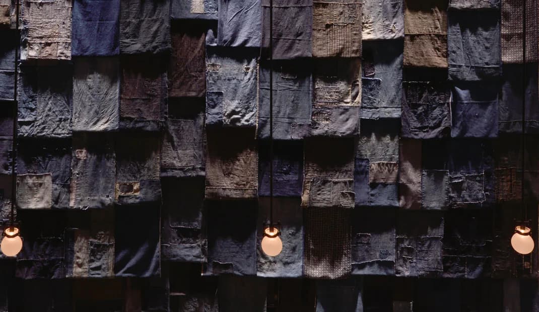 Dark textured wall installation with stacked slate or stone tiles in varying shades of gray and blue, punctuated by three warm-glowing pendant lights, by Osamu Tsuchiya.