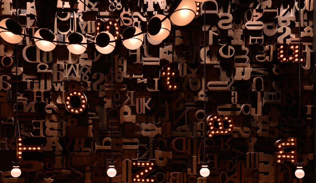 Dark installation with suspended copper-colored metal letterforms and illuminated signage creating an abstract typographic environment by Osamu Tsuchiya.