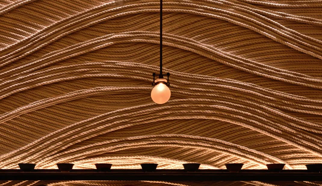 Curved wooden ceiling with linear grain patterns and a minimalist pendant light fixture, creating warm architectural detail by Osamu Tsuchiya.