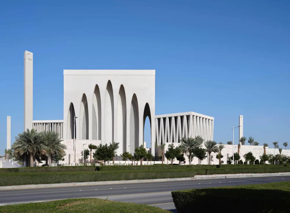 White modernist mosque with dramatic arched openings, surrounded by palm trees and landscaping, designed by Osman Chaudhry.