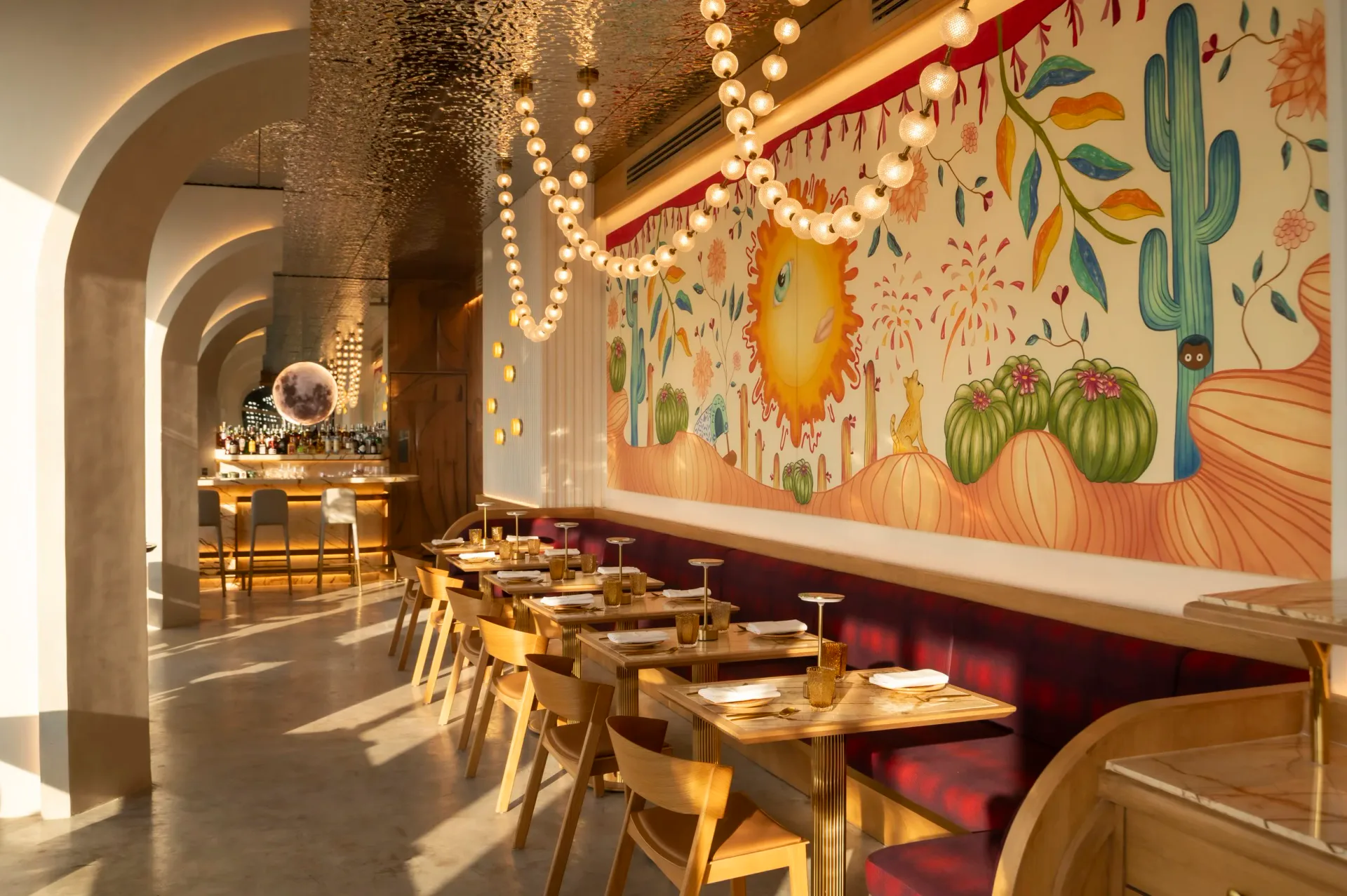Warm-toned restaurant interior with arched brick ceiling, string lighting, wooden chairs, red booth seating, and whimsical mural artwork featuring cacti and sun motifs by Pablo Castellanos.