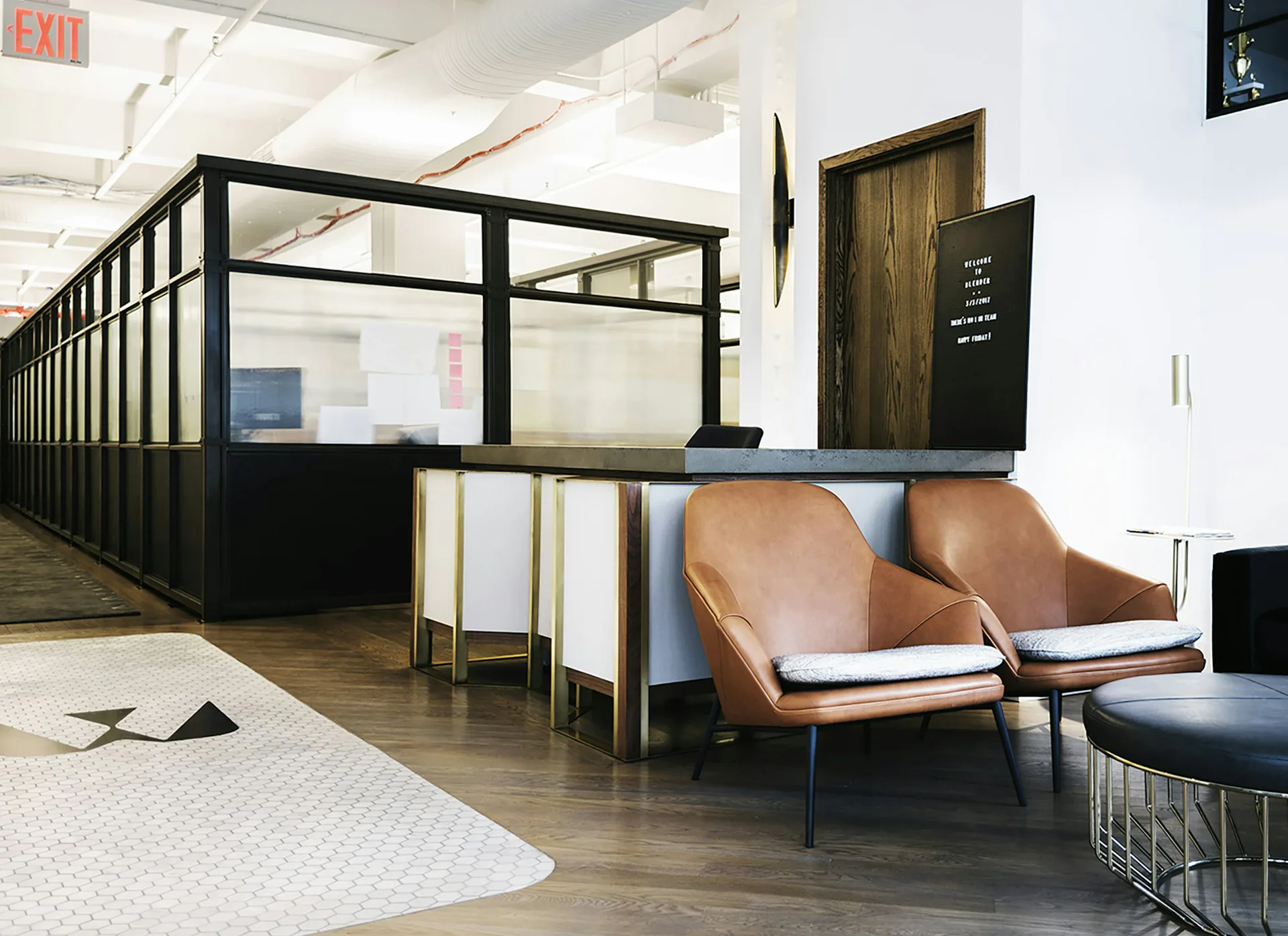Modern office reception area featuring black-framed glass partitions, cognac leather lounge chairs, wooden flooring, and minimalist furnishings by Pam Chaiyachakorn.