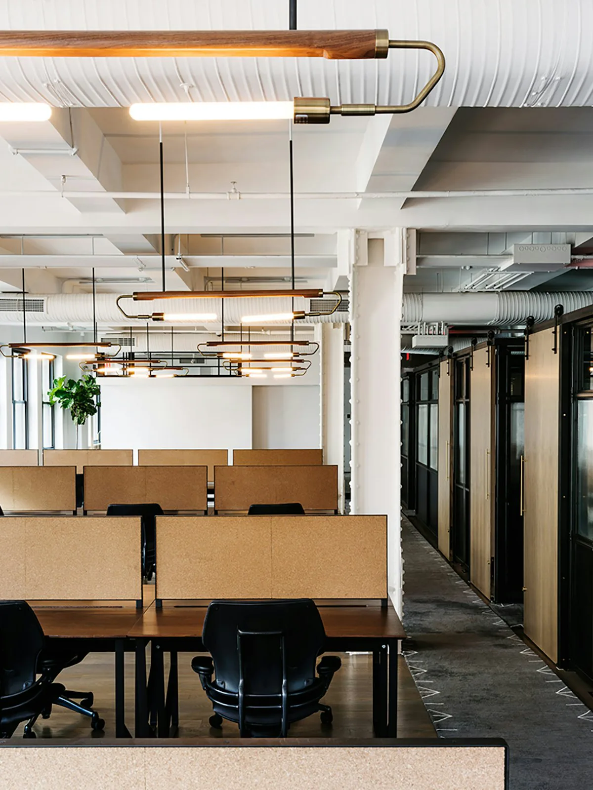Modern office interior featuring natural wood workstations, brass pendant lighting, white coffered ceiling, and dark corridor with glass partitions by Pam Chaiyachakorn.