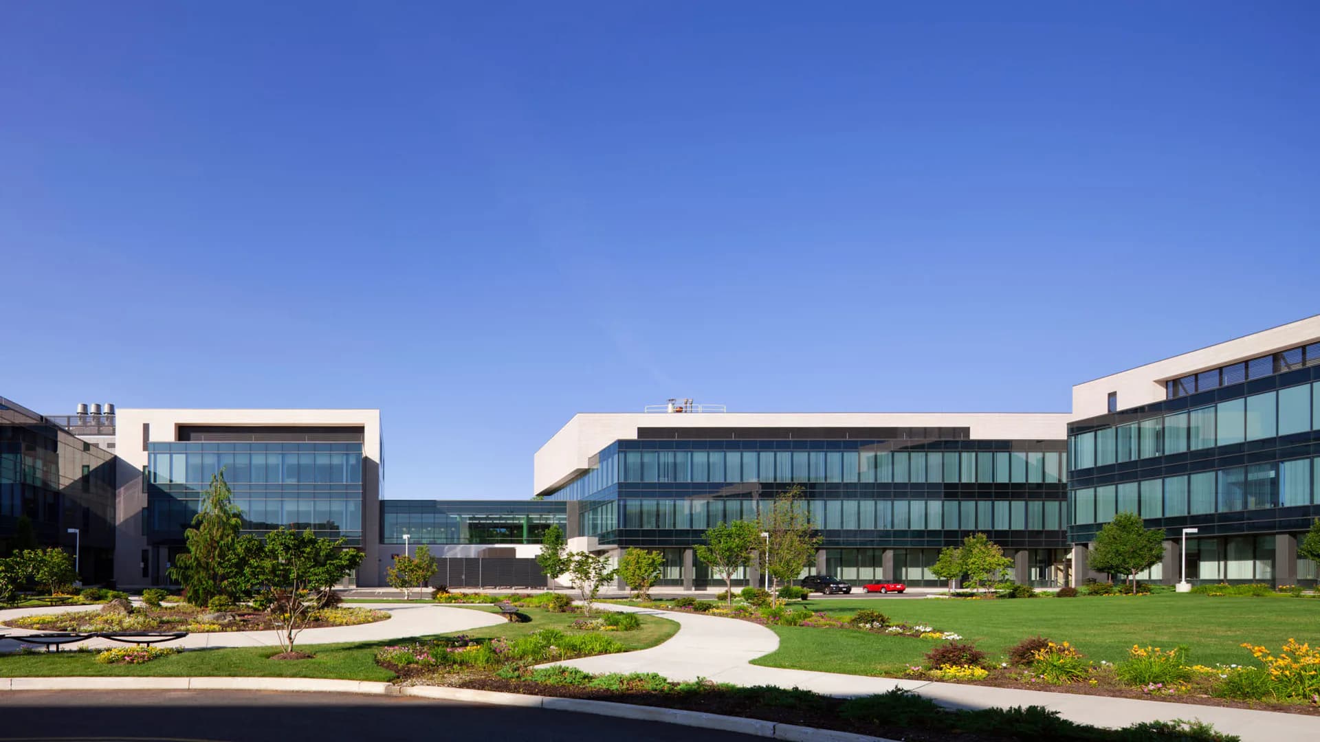 Modern institutional campus with curved glass and metal buildings, manicured lawns, and contemporary landscaping featuring geometric pathways and flowering gardens by Pamela Cole.