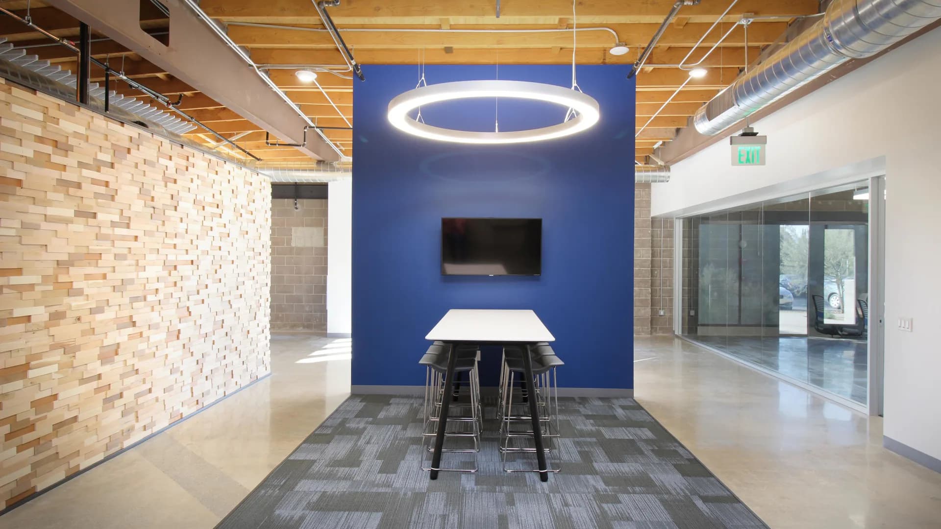 Modern office break room with bold blue accent wall, circular ceiling light, wooden brick feature wall, and high-top table seating by Pamela Cole.