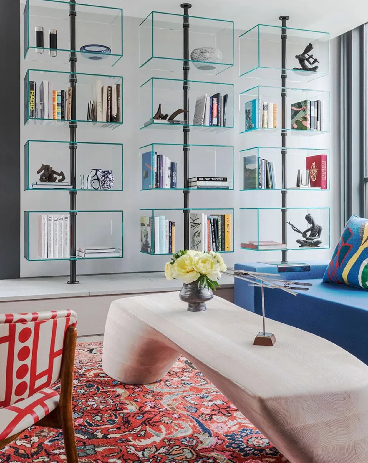 Modern living room with floor-to-ceiling glass and metal shelving displaying books and objects, blue upholstered bench, patterned rug, and white sculptural seating by Pamela R Ackerman.